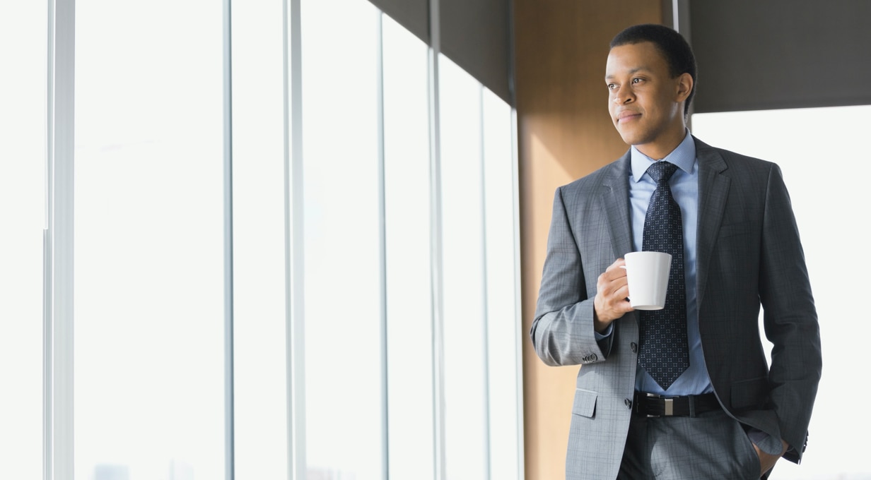 A businessperson holding a coffee cup looks out the windows of an office.