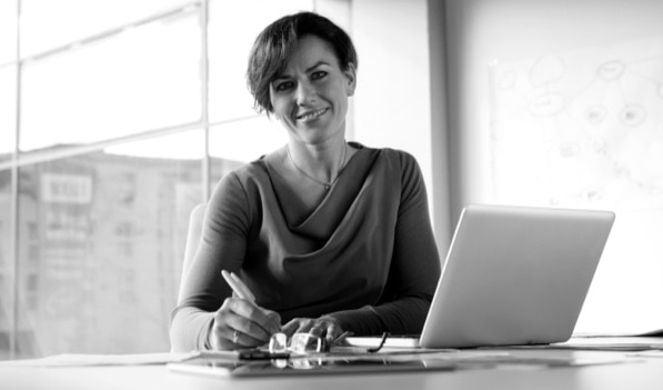 A smiling businessperson sits at a desk with a computer and writes down notes.