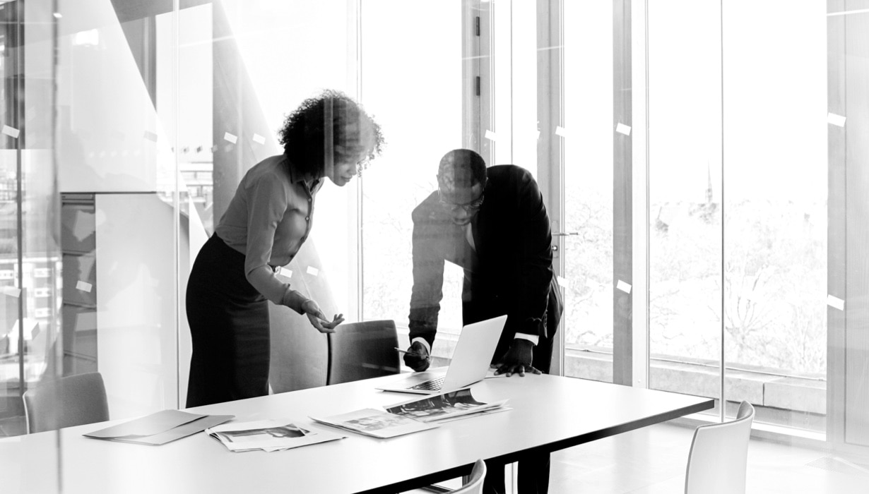 Two businesspeople stand at a conference table and work together at a laptop.