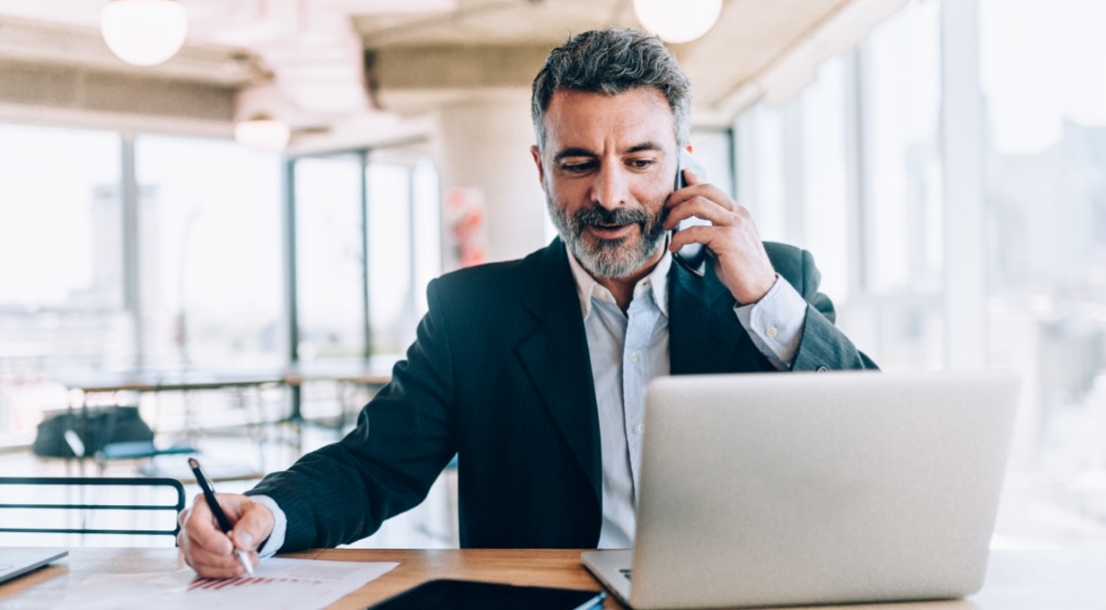 A businessperson talks on a cell phone while looking at a laptop and taking notes on paper.