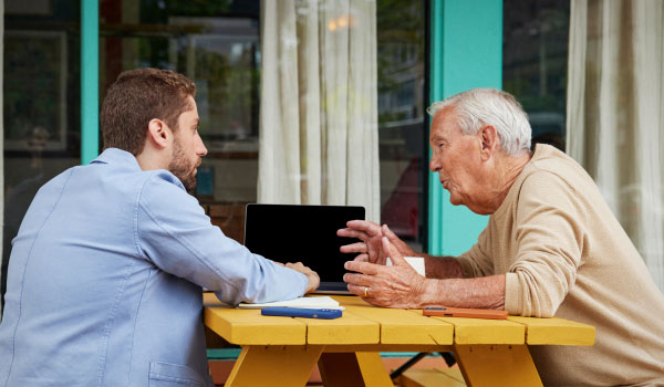 Older man and younger man talking at a picnic table with a laptop.