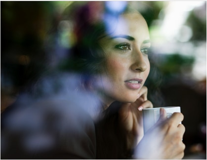 A woman drinks coffee while pensively looking out a window.